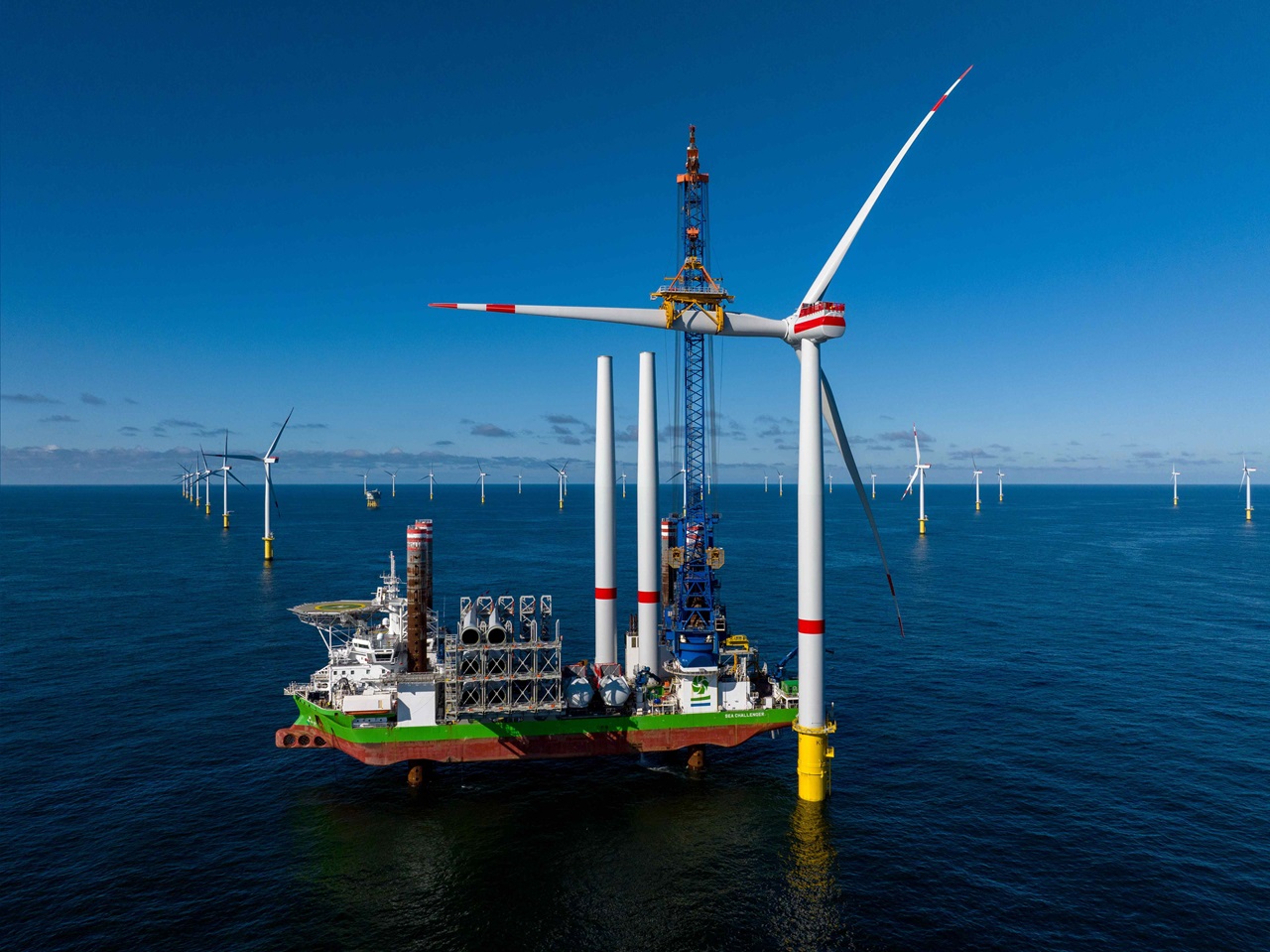 A large offshore wind turbine installation vessel with multiple wind turbines in the background under a clear blue sky.