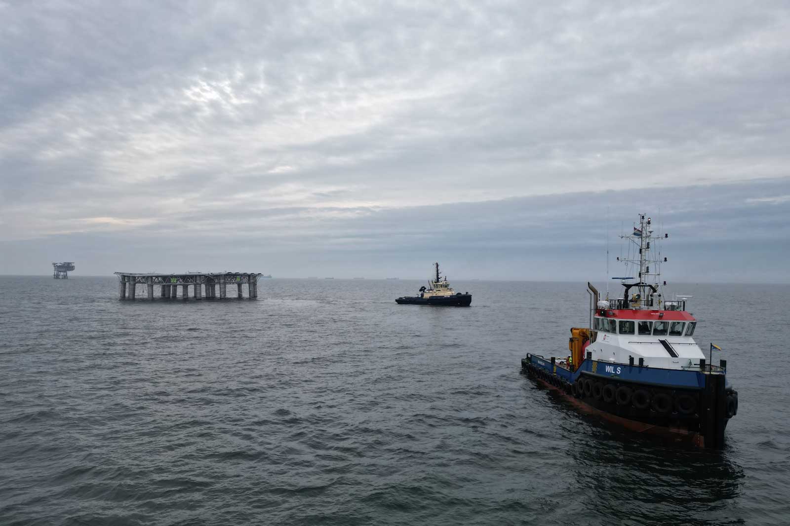 Zwei Schlepperboote navigieren im Meer, während eine Plattform im Hintergrund sichtbar ist und der Himmel bewölkt ist.