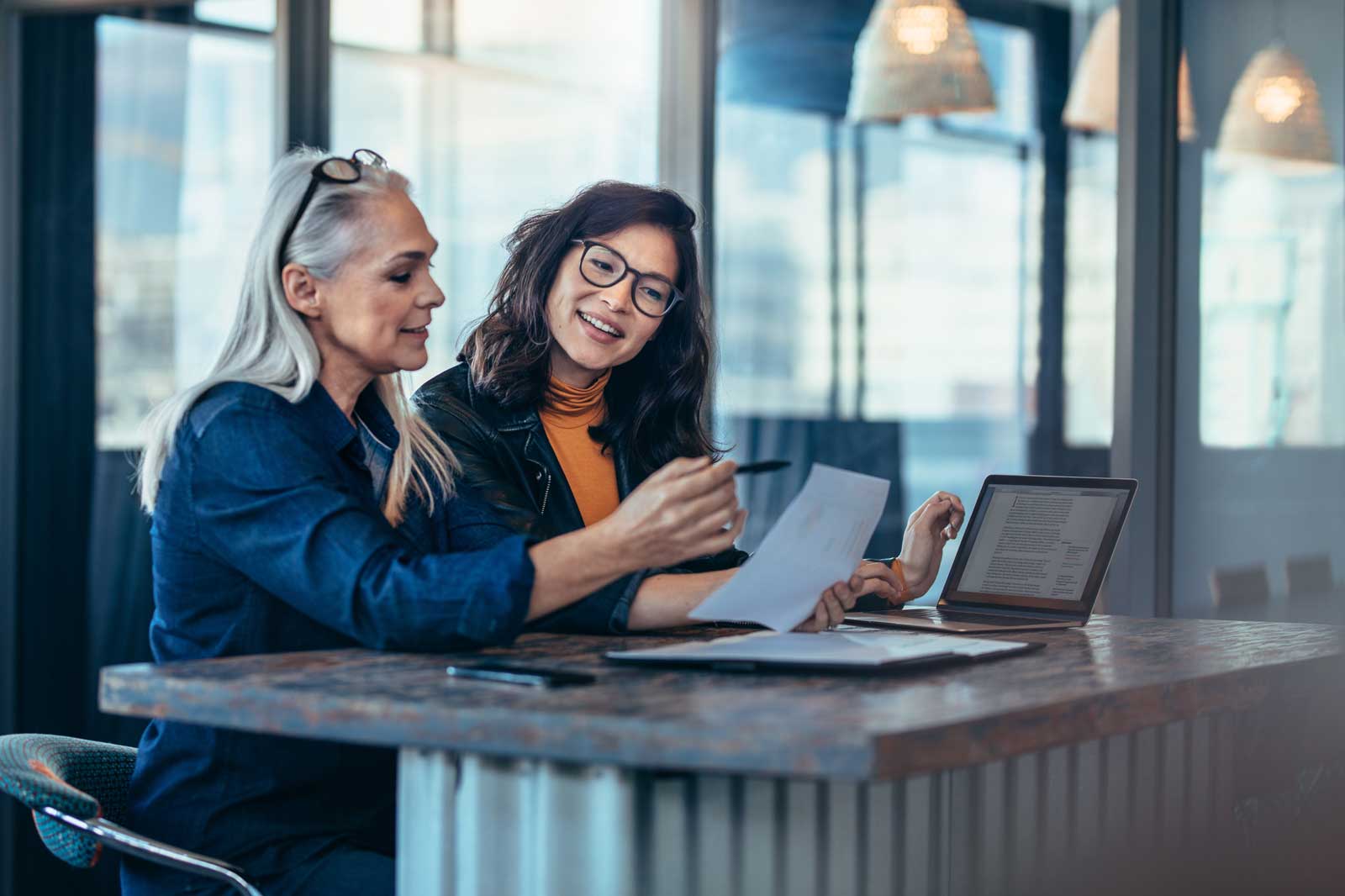 Zwei Frauen sitzen an einem Tisch und diskutieren, während sie Dokumente und einen Laptop benutzen.