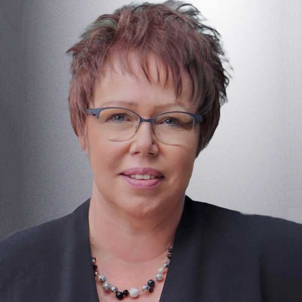 Eine Person mit kurzem, braunem Haar trägt eine schwarze Bluse und eine Halskette mit verschiedenen Perlen. Portrait of Ulrike Pugh.