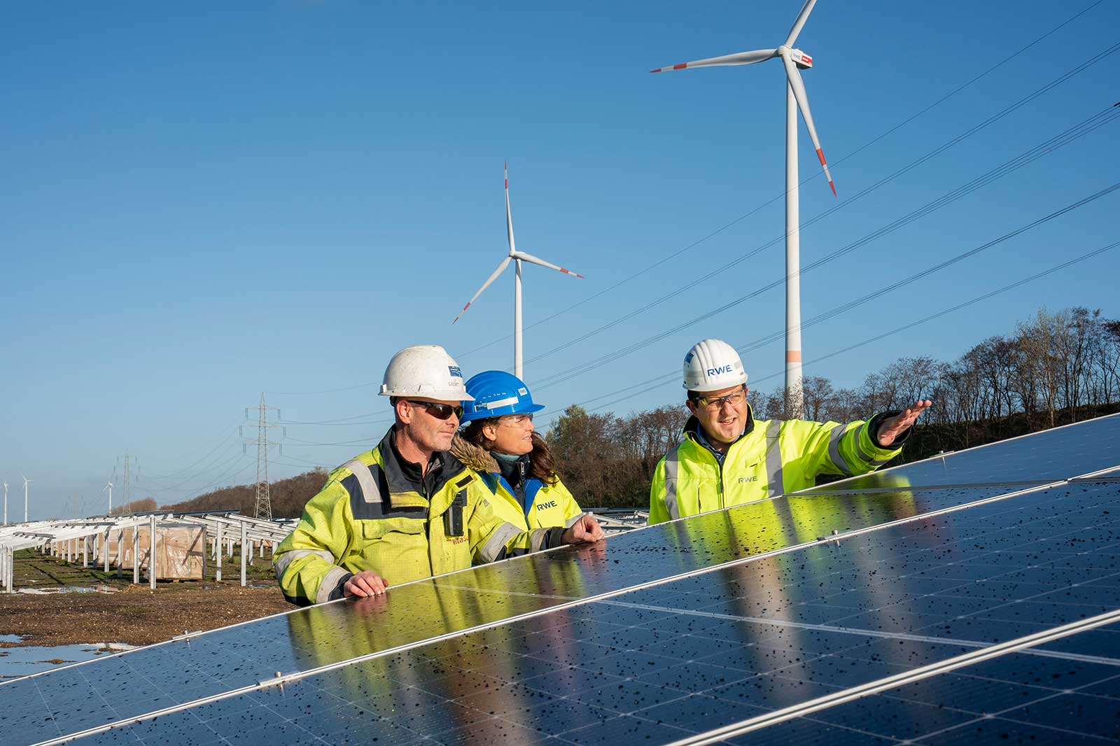 Fachleute inspizieren Solarpanel mit Windkraftanlagen im Hintergrund unter blauem Himmel.