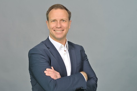 Ein Mann in einem dunklen Anzug mit verschränkten Armen, vor einem grauen Hintergrund. Er trägt ein weißes Hemd. Portrait von Götz Hanau.