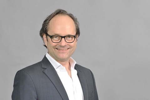 Eine Person in einem dunklen Anzug und einem weißen Hemd posiert vor einem schlichten grauen Hintergrund. Portrait von Henning Joswig.