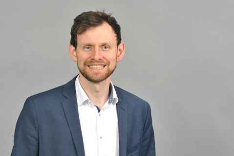 Ein Mann trägt einen Anzug und ein Hemd, steht vor einem grauen Hintergrund. Portrait von Jan Josef Bernholz.