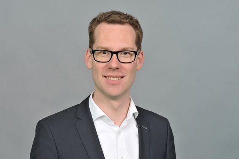 Ein Mann trägt einen schwarzen Anzug und ein weißes Hemd. Der Hintergrund ist grau. Portrait von Kevin Pasch.