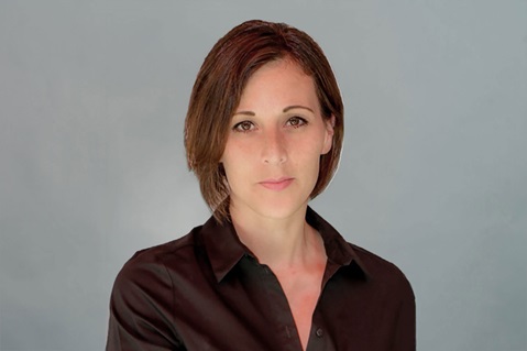 Eine Person mit schulterlangen Haaren trägt ein schwarzes Hemd und steht vor einem grauen Hintergrund. Portrait von Natalie Henßen.