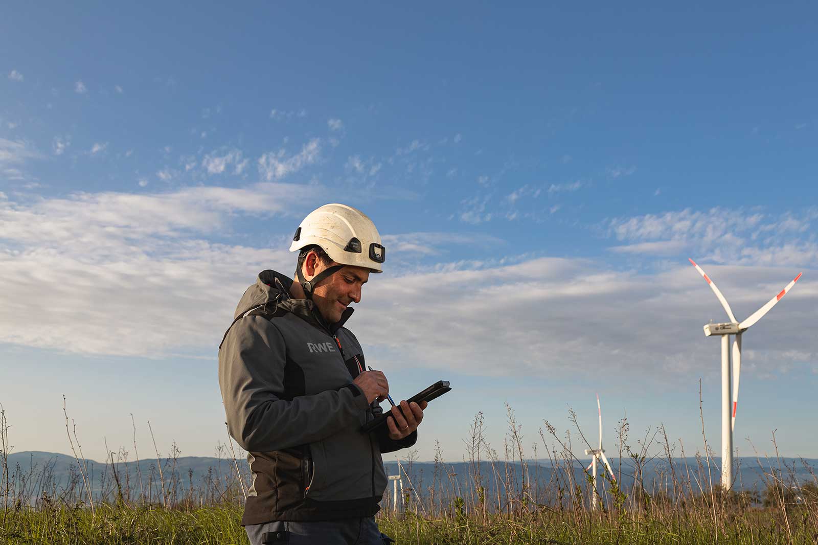 A worker in a safety helmet uses a mobile device at a wind farm under a clear sky.