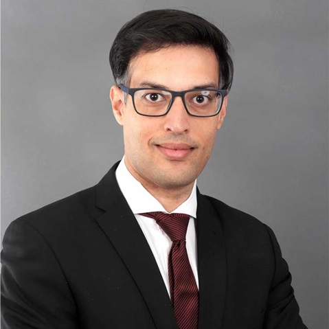 Ein Mann in einem schwarzen Anzug und rotem Binder vor einem grauen Hintergrund, der eine professionelle Haltung einnimmt. Portrait von Kunal Chandra.