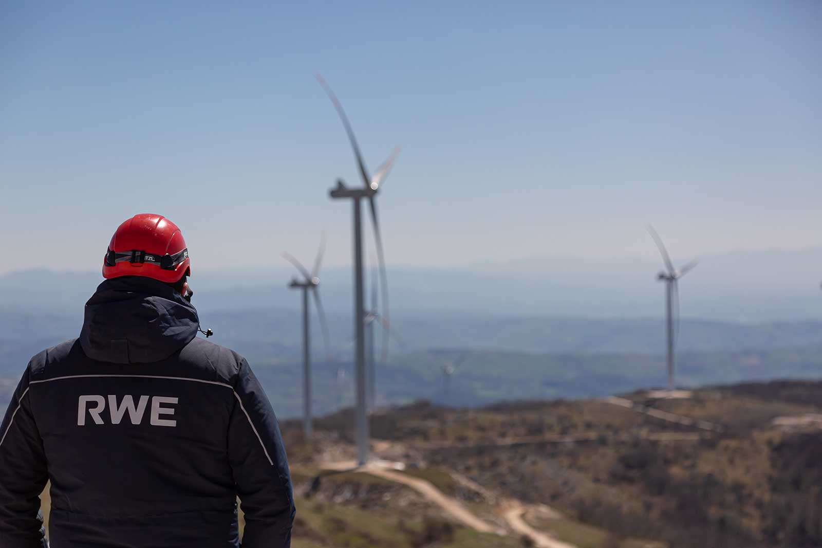 Ein Mitarbeiter mit rotem Helm steht an einem Windpark und beobachtet die Windturbinen in der Ferne.