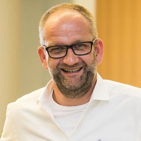 Ein Mann trägt ein weißes Hemd und ist in einem modernen Raum abgebildet, mit schlichten Holzwänden im Hintergrund.