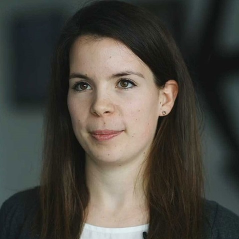 Eine Person mit langen, dunklen Haaren trägt ein graues Oberteil und Ohrringe. Der Hintergrund ist unscharf.