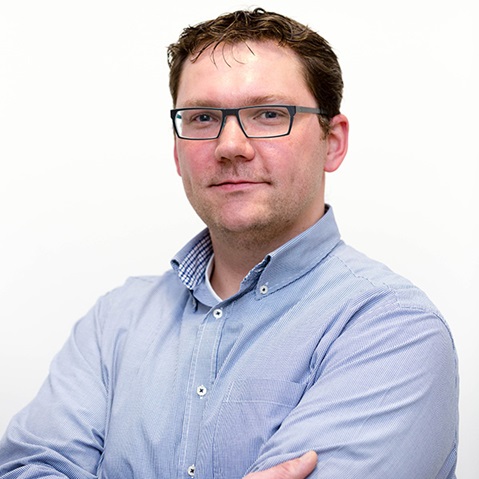 Ein Mann mit verschränkten Armen trägt ein kariertes Hemd und blickt zur Kamera, vor einem weißen Hintergrund.