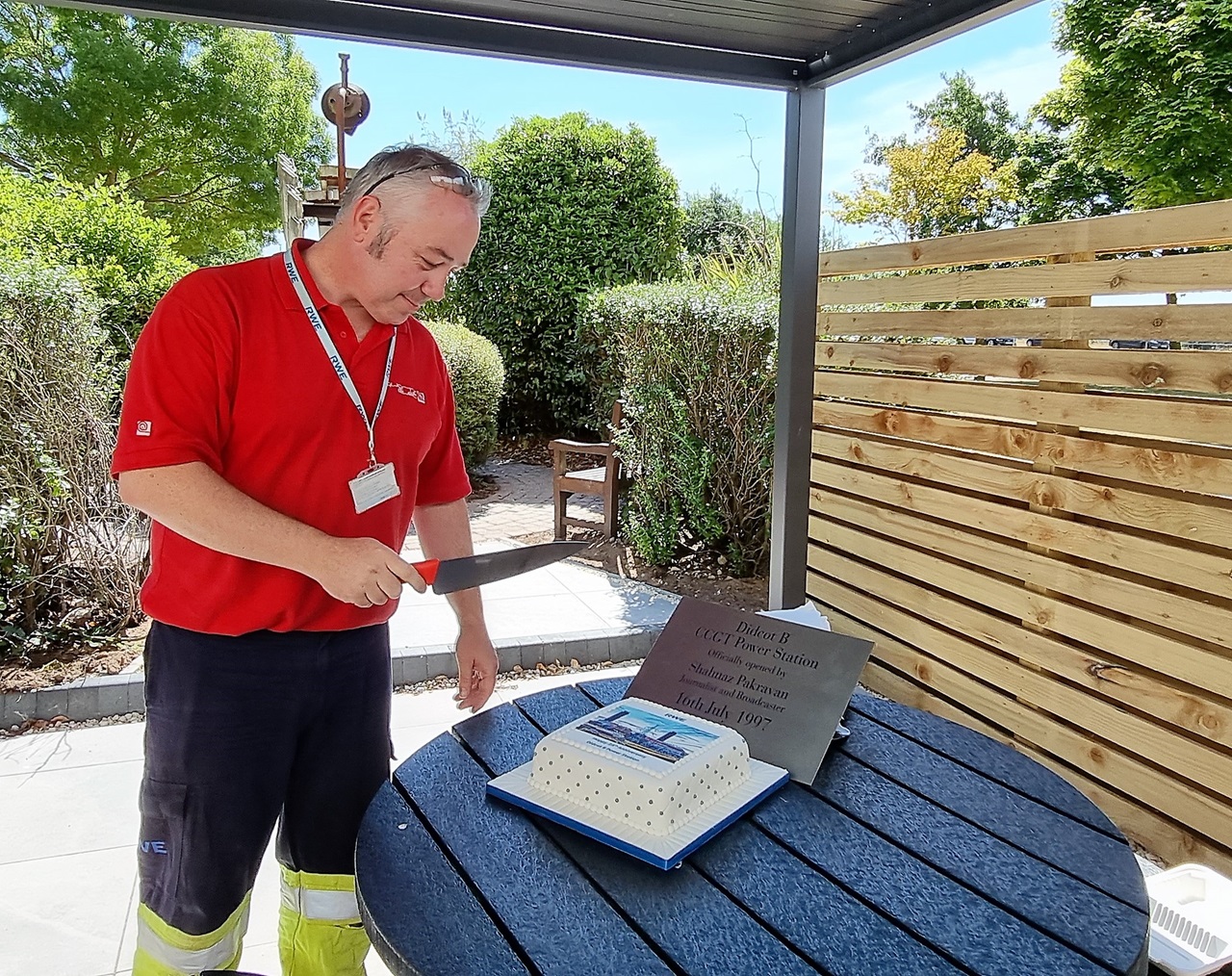 A man in a red shirt is cutting a birthday cake on an outdoor terrace. Greenery and a wooden fence are in the background.