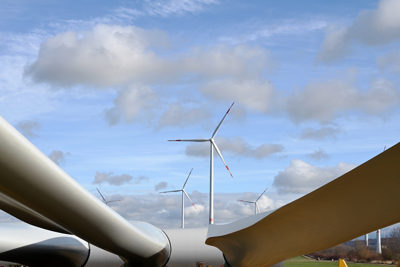 Wind turbines in the background framed by the blades of a wind turbine in the foreground against a blue sky with clouds.