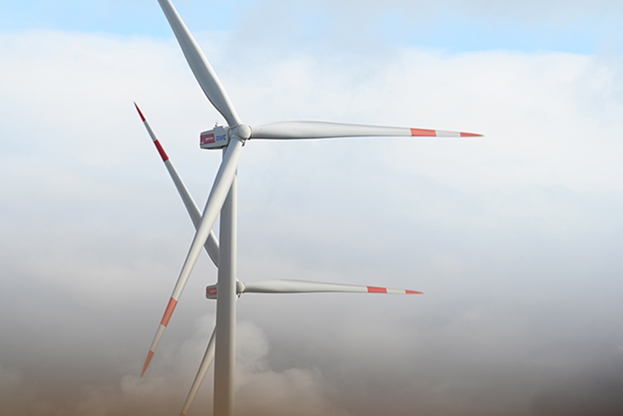 Three wind turbines with red and white markings, partially obscured by clouds, against a clear blue sky.
