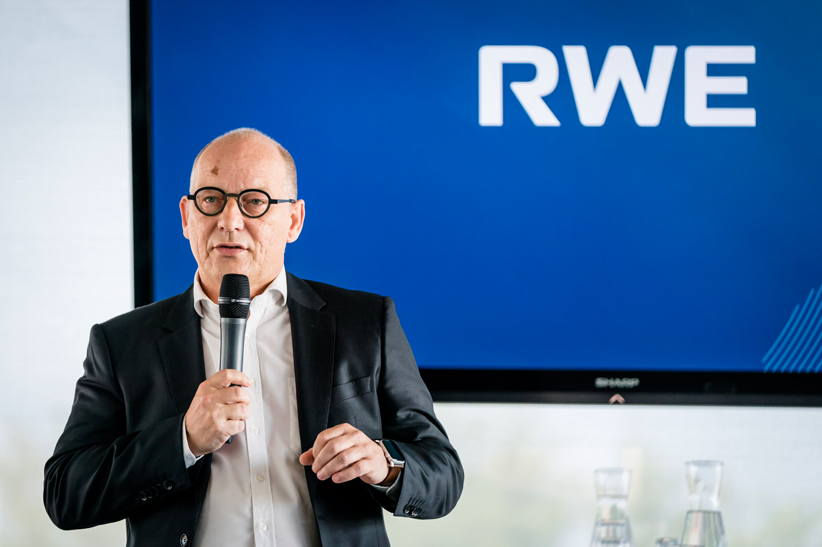 A speaker in a black suit holds a microphone in front of a large screen displaying the RWE logo.