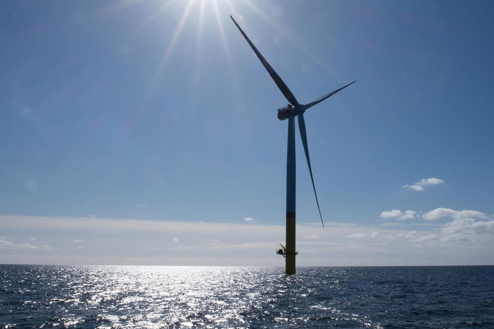 A large wind turbine stands tall against a clear blue sky, with the sun shining brightly over sparkling ocean waters.