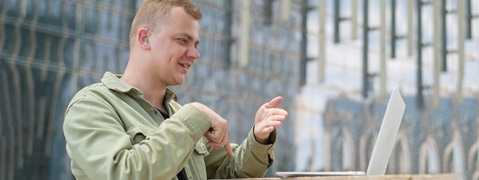 A person seated outdoors, gesturing while using a laptop in front of a modern, glass building.