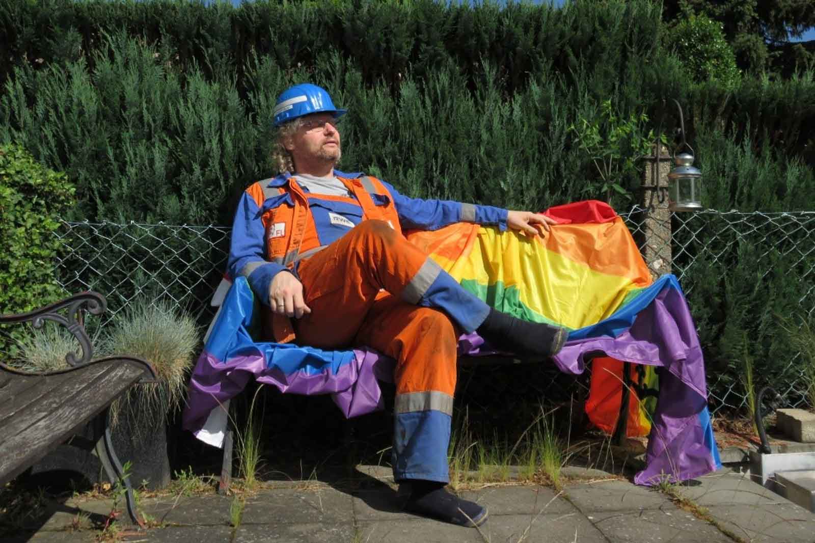 Ein Arbeiter in orangefarbener Latzhose sitzt auf einer Bank, die mit einer Regenbogenflagge geschmückt ist, umgeben von Grün.