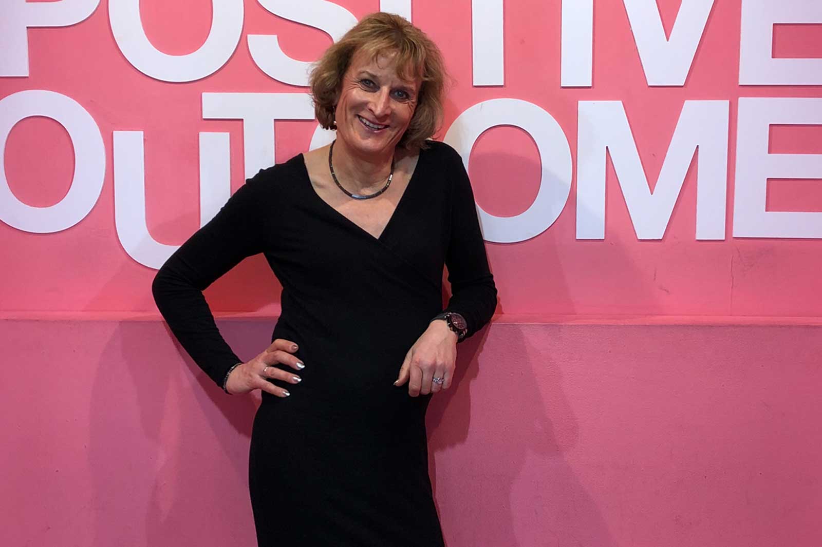 Eine Person in einem schwarzen Kleid steht vor einer rosa Wand mit dem Text 'POSITIVE OUTCOME'.