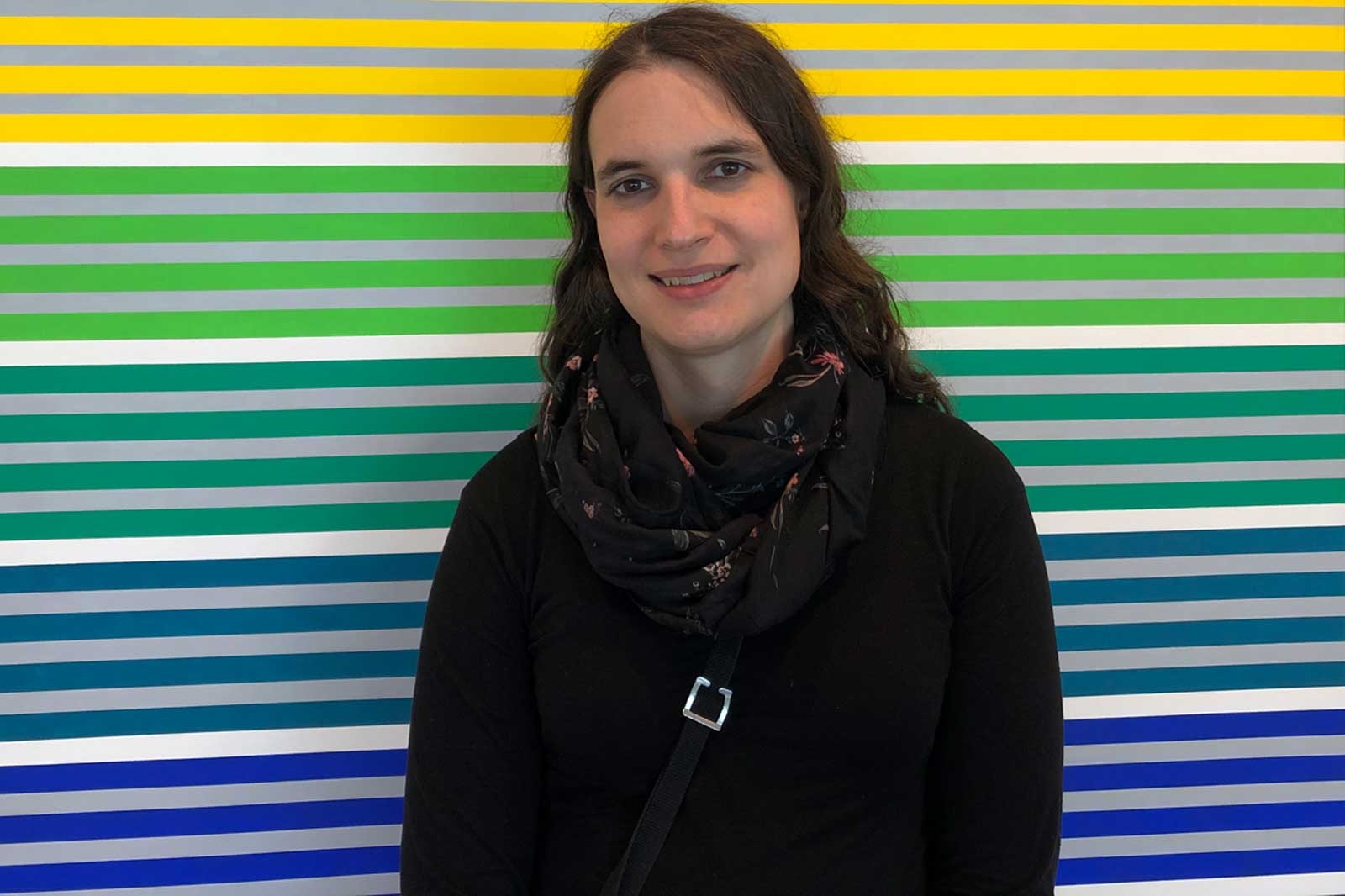 A person stands in front of a colourful striped background in various hues, wearing a black top and a scarf.