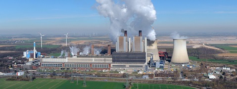 An industrial complex featuring smokestacks, cooling towers, and wind turbines against a rural backdrop.