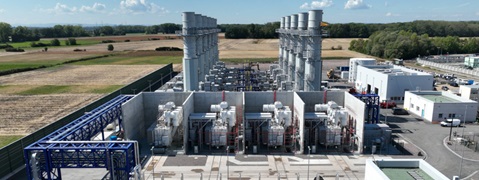 A power plant featuring large turbines and transformers, surrounded by fields and a clear blue sky.
