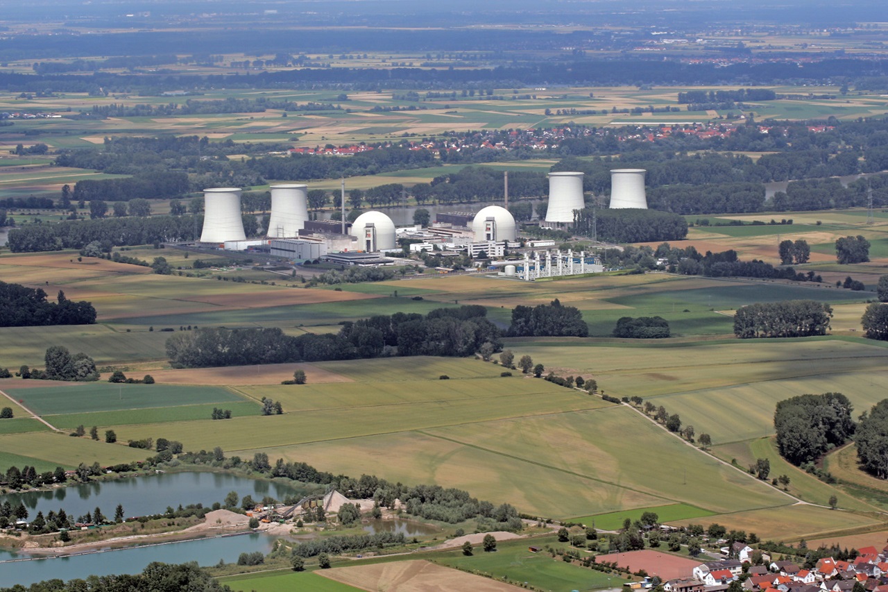 Aerial view of a nuclear power plant surrounded by fields and a small lake. Cooling towers and several buildings are visible.