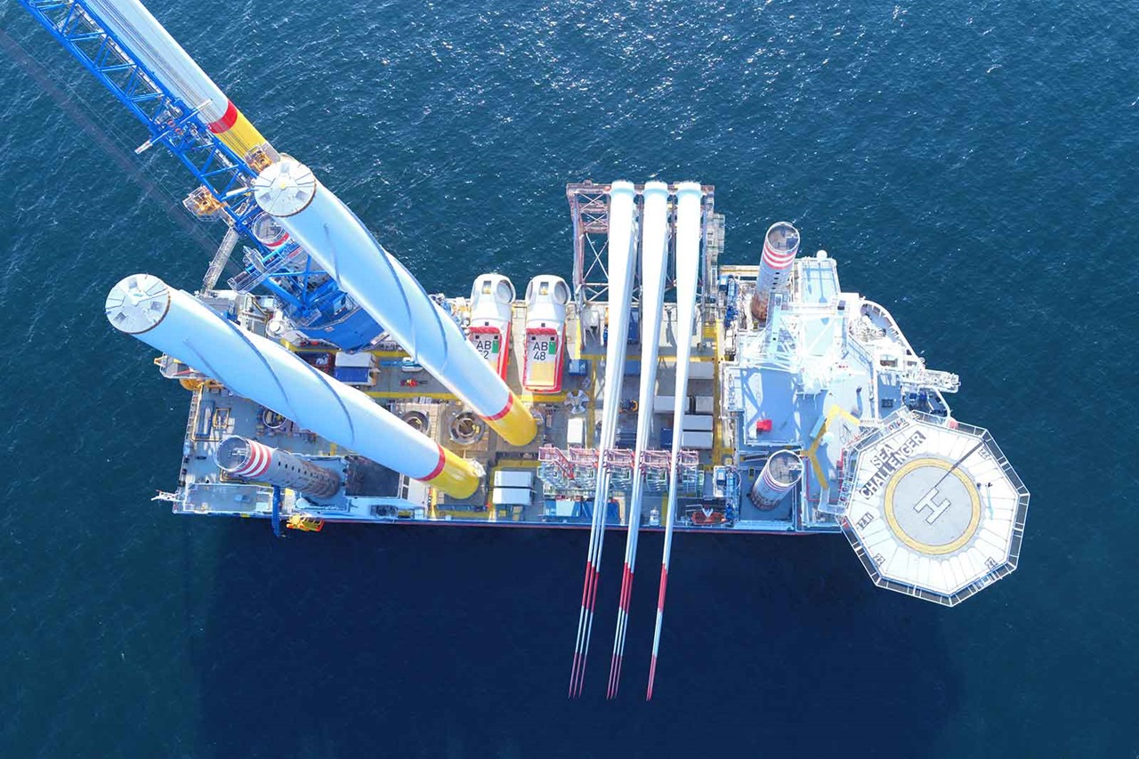 An aerial view of a large offshore vessel with wind turbine blades and a crane on deck, surrounded by deep blue water.