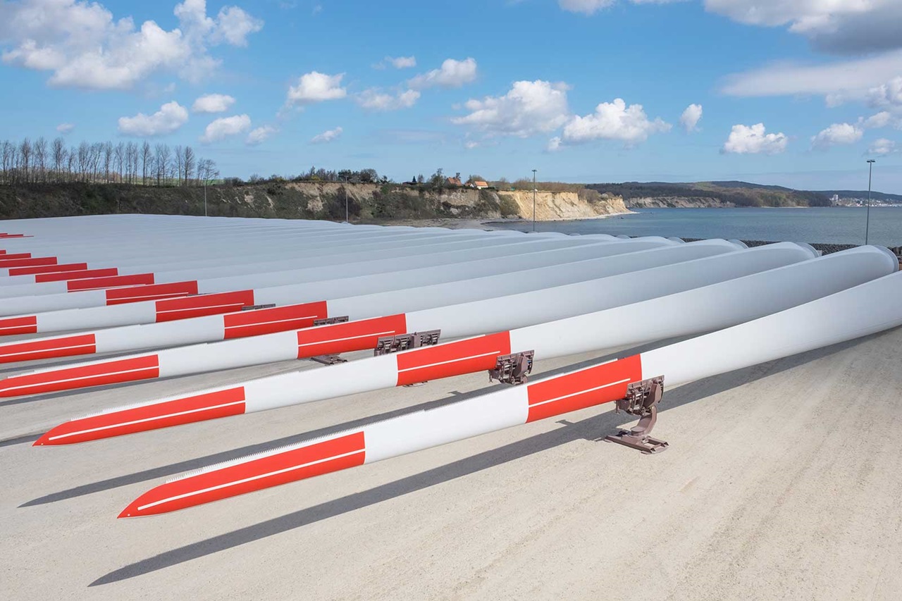 A row of wind turbine blades with red and white stripes, resting on a concrete surface near the coast.