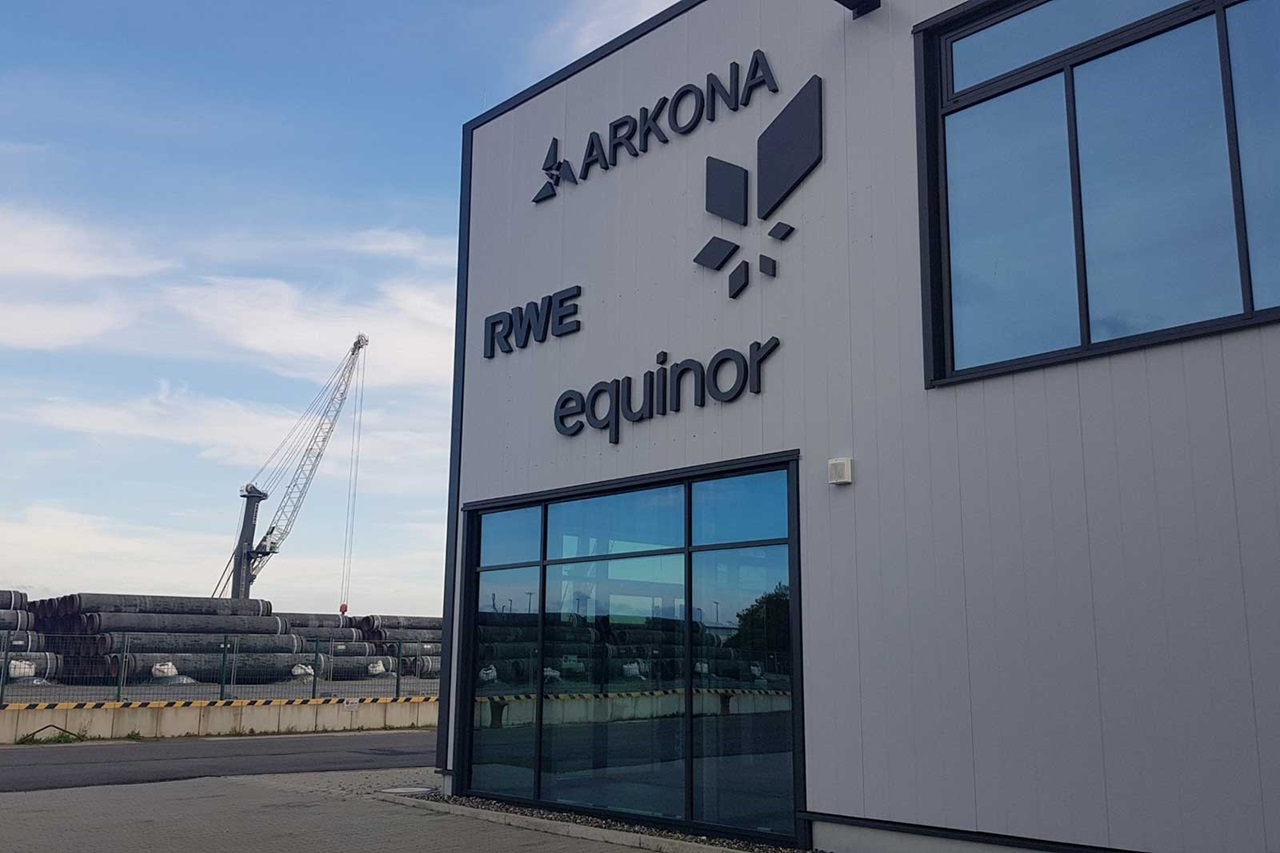 The building's exterior features the logos of Arkona, RWE, and Equinor. Stacked pipes and a crane are visible in the background.