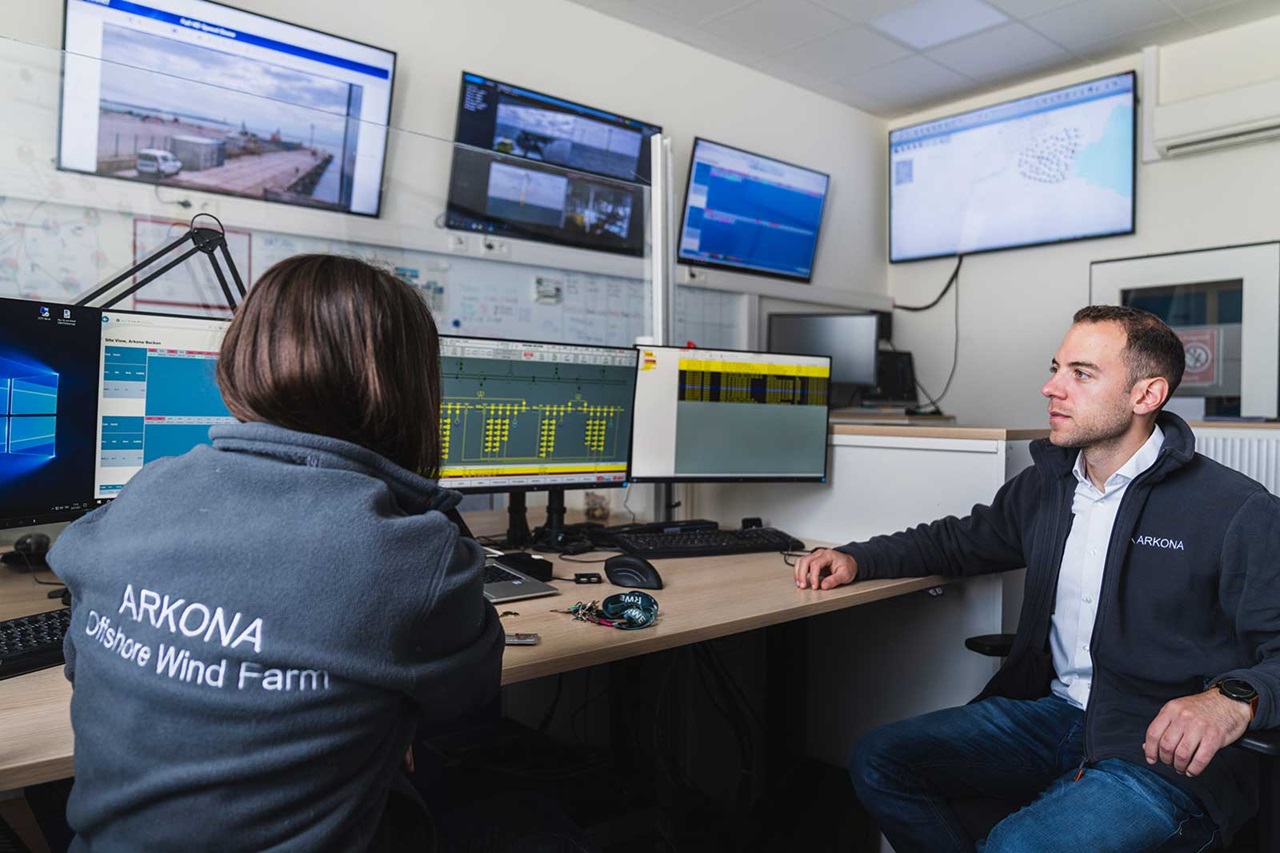 Two individuals in the control room of an offshore wind farm, surrounded by monitors and technical equipment.
