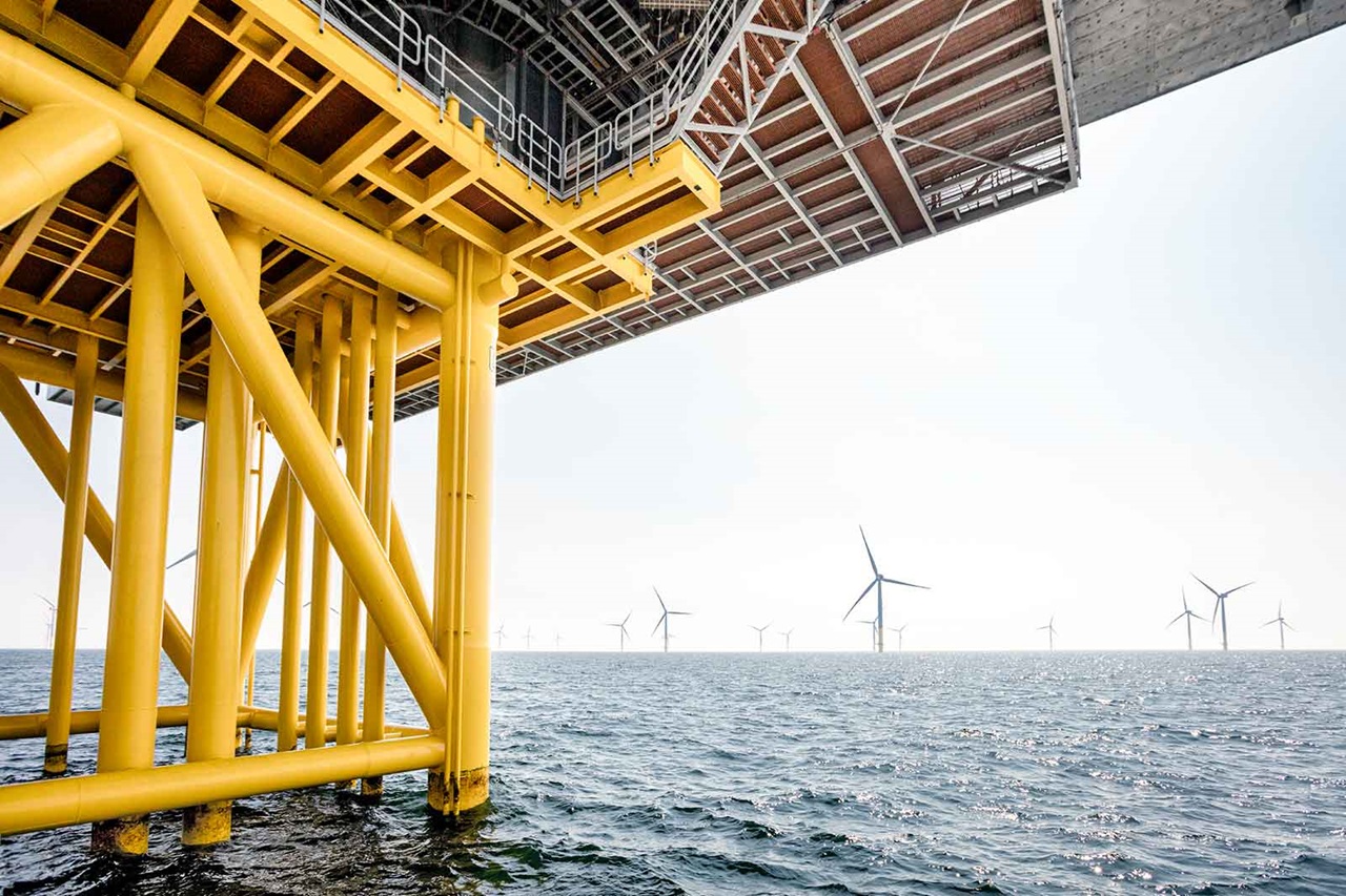 A view of the yellow structure of an offshore installation, with wind turbines in the background over the water.