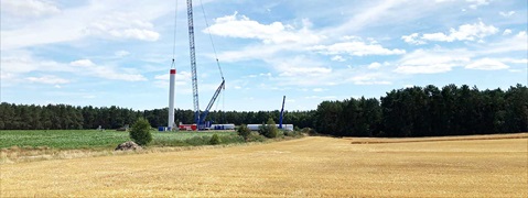A wind turbine is being erected on a field with a crane. Forests and blue skies are visible in the background.