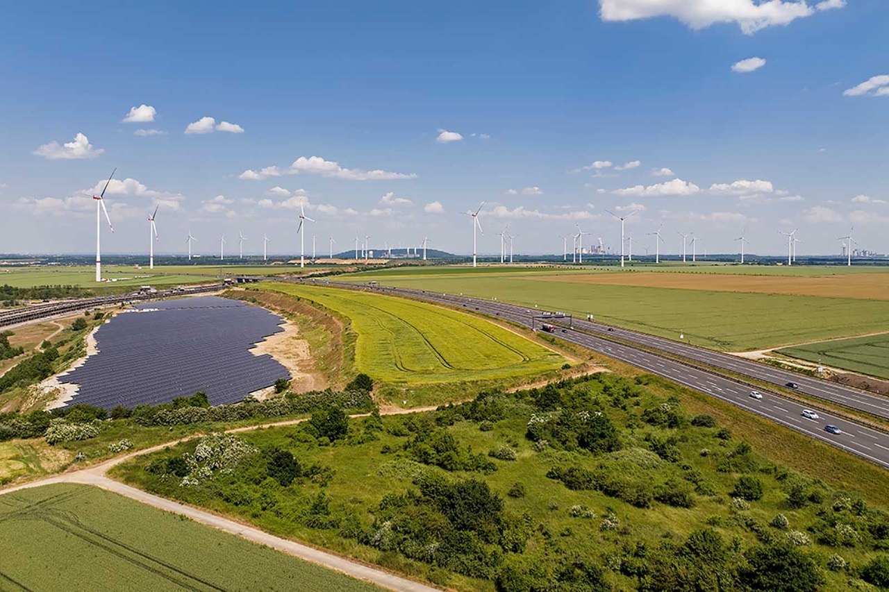 Landscape with wind turbines and solar panels, green fields, and a road under a blue sky.