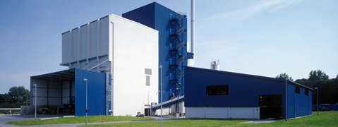 A modern industrial building with a blue facade, large windows, and a tall chimney against a clear sky.
