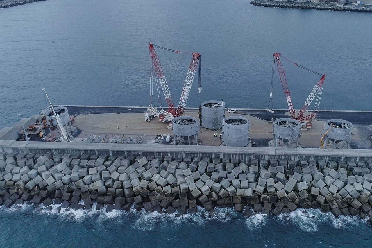 View of a harbour facility with cranes, cylinders, and a concrete wall by the water.