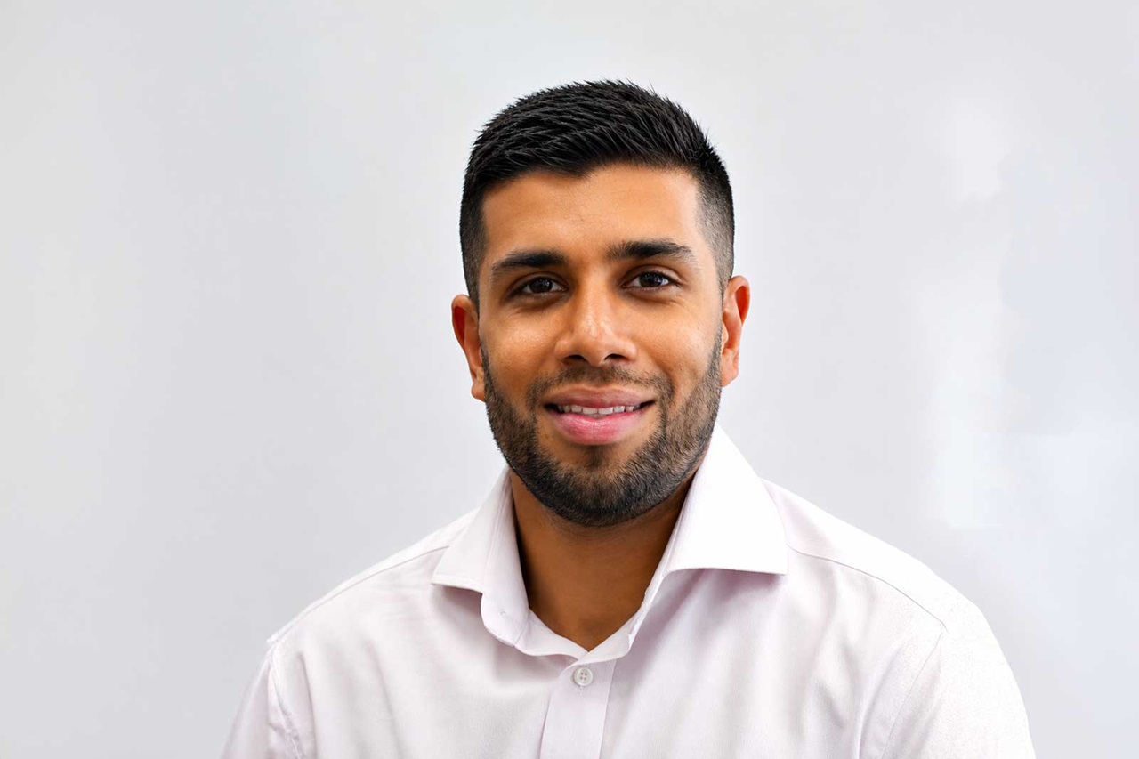 A young man with short dark hair and a trimmed beard smiles warmly while wearing a white collared shirt against a pale gray background.Portrait of Akhil Shah.