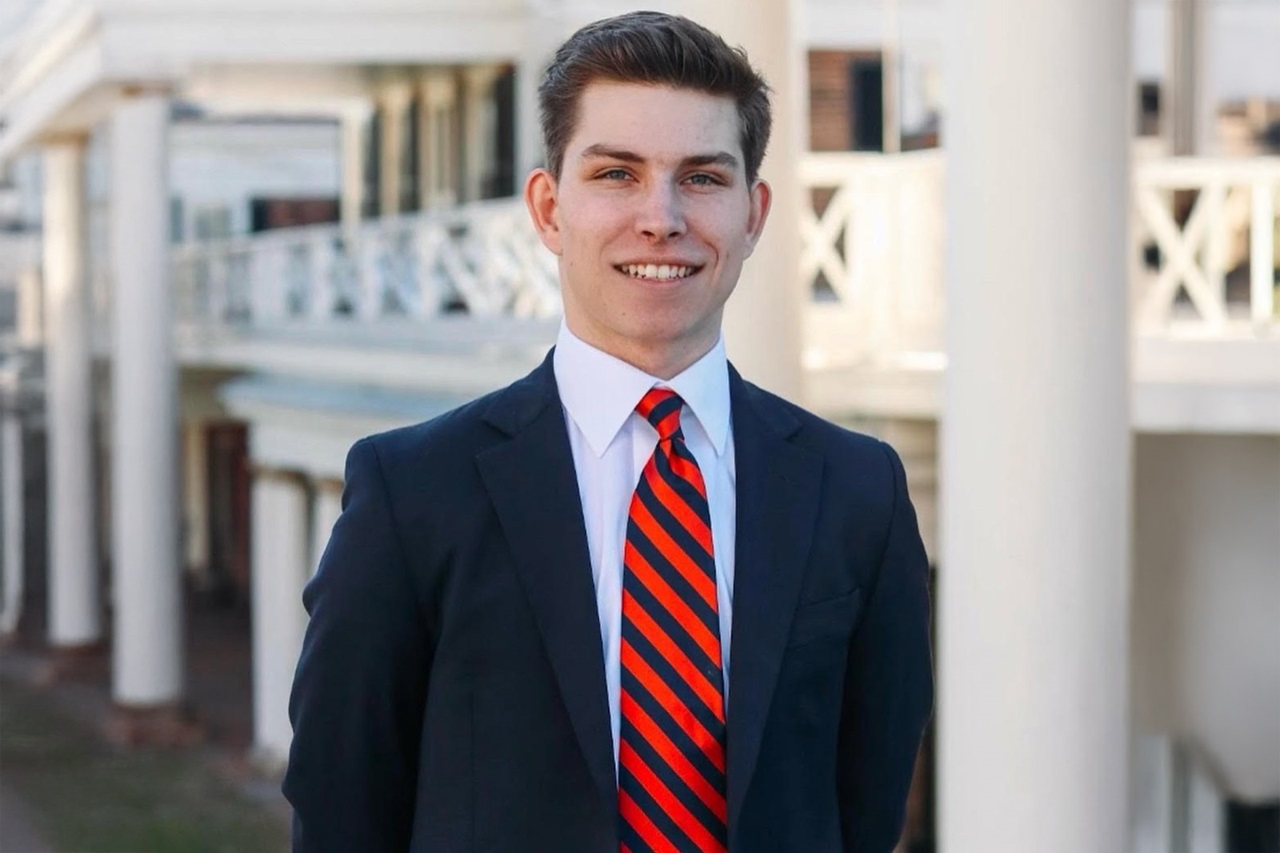 A man in a black suit and red-striped tie stands in front of a building with white railings. Portrait of Bill Cull.