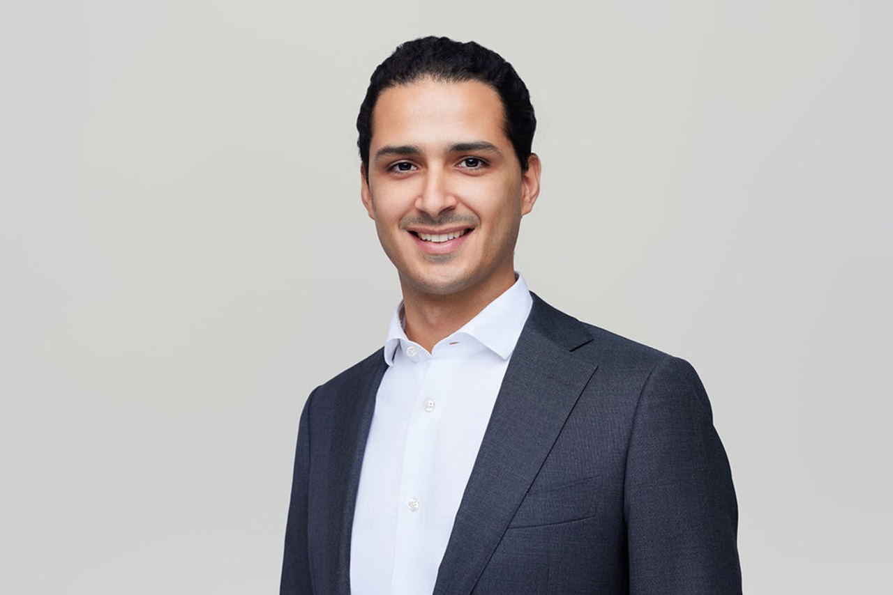 A man in a dark suit with a white shirt stands against a plain light background. Portrait of Marwan Essabar.
