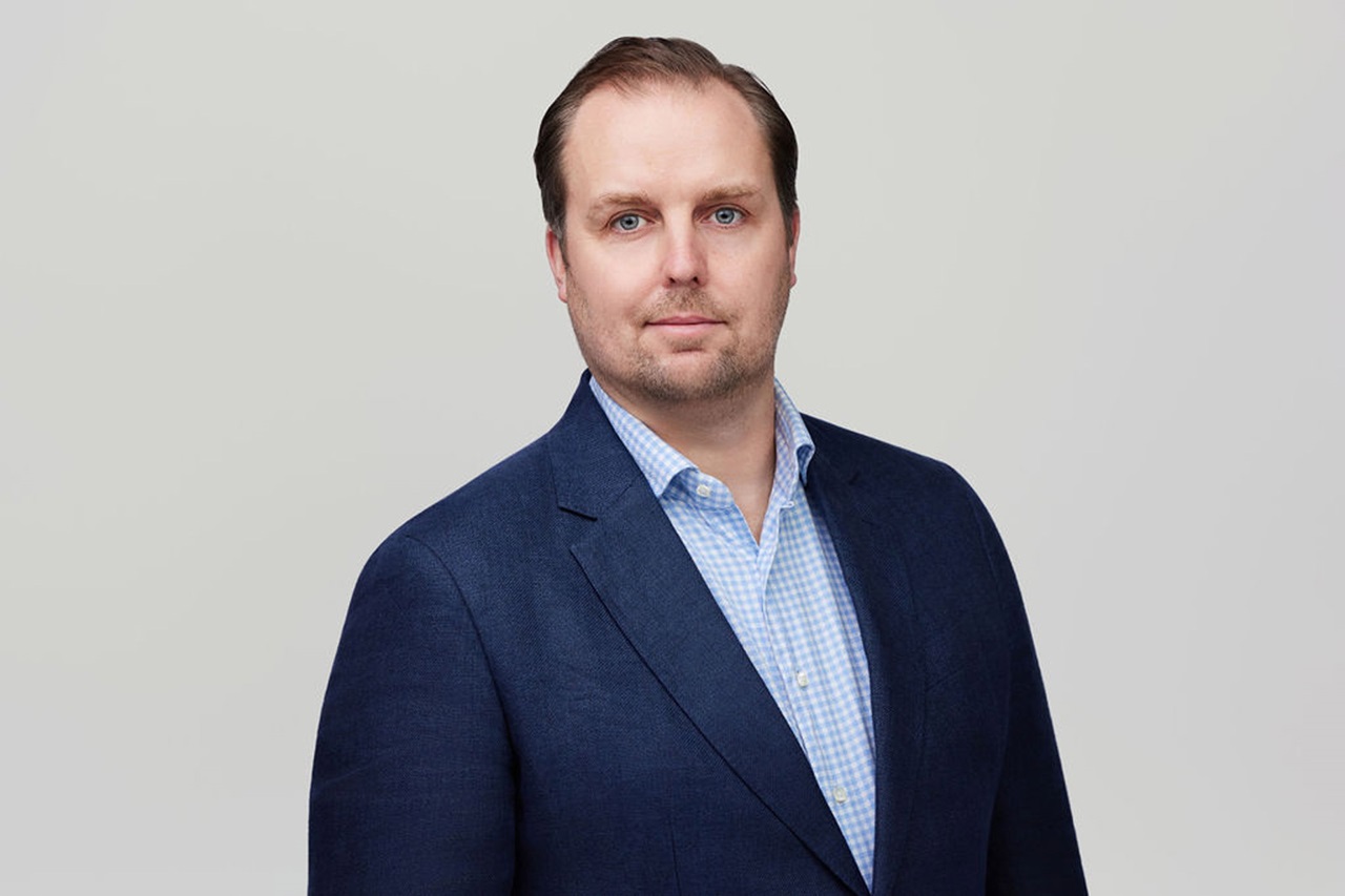 A man in a navy blazer over a light blue checkered shirt stands confidently against a plain background. Portrait of Rob MacIntyre.