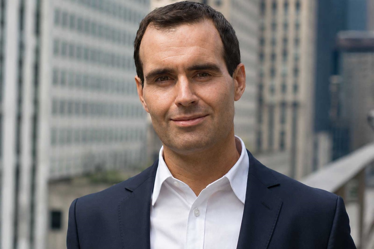 A man in a dark navy suit and white shirt stands on a rooftop terrace with gray skyscrapers behind him, wearing a slight smile. Portrait of Stephen O'Reilly.