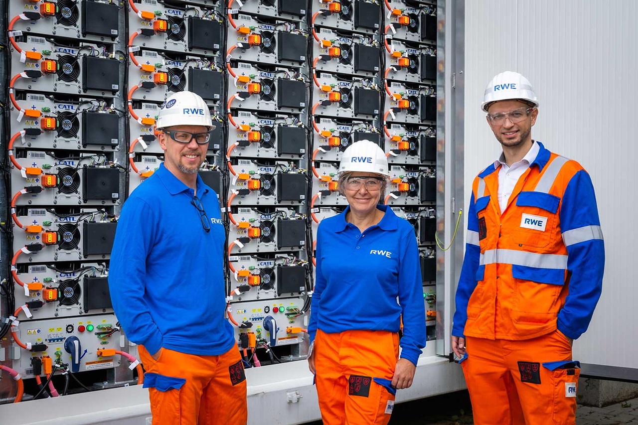 Three workers in blue and orange overalls stand in front of a wall with battery modules and electrical components.