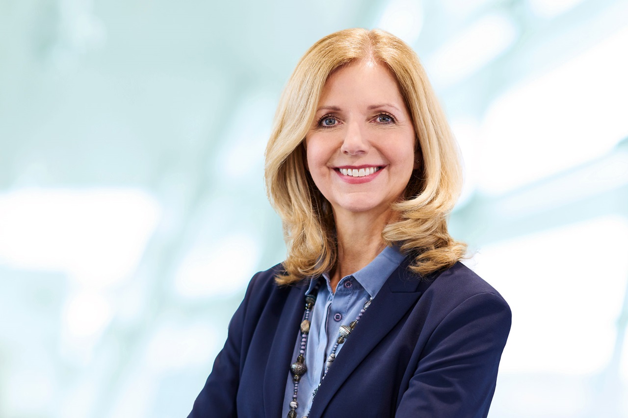 A woman with wavy blonde hair is wearing a blue suit and a blue blouse. The background is bright and blurred. Portrait of Katja van Doren.