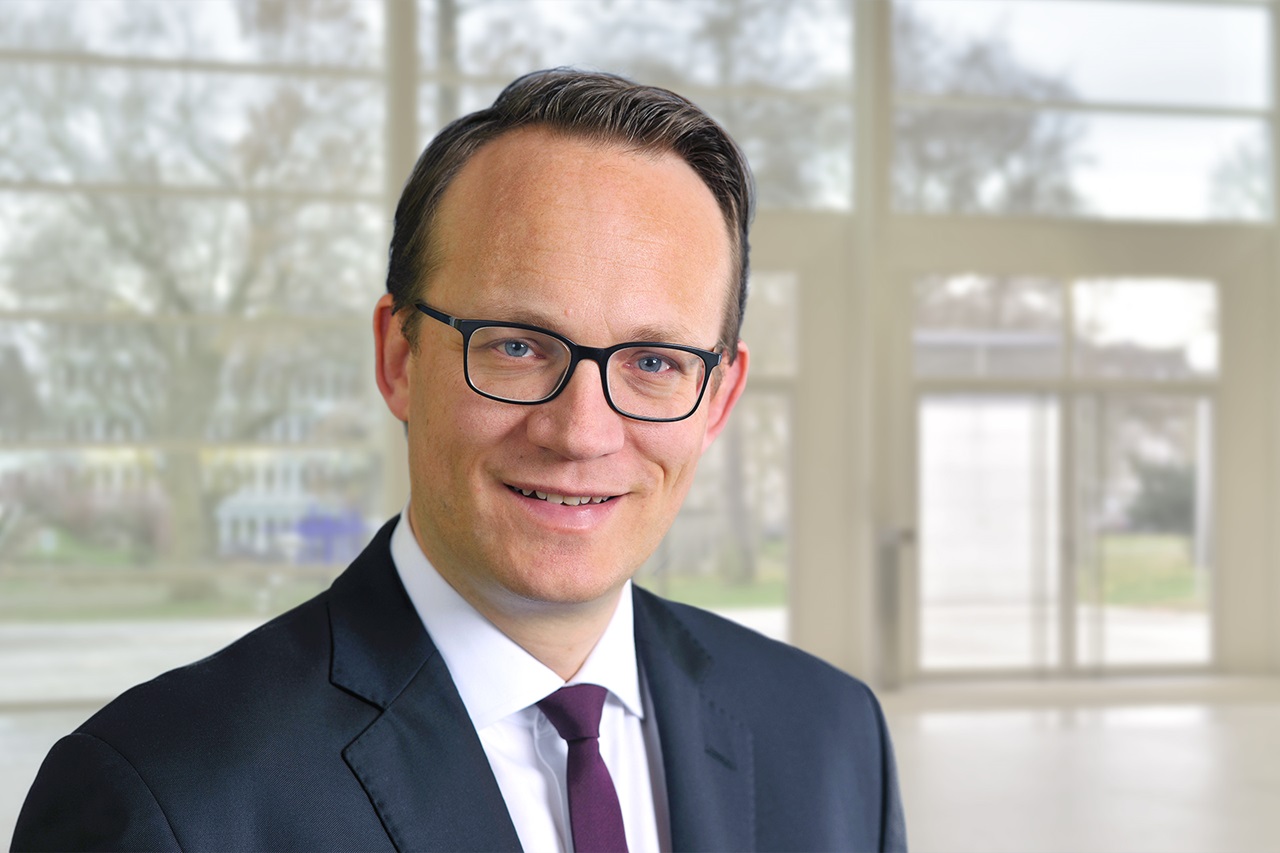 A man in a suit stands in front of a modern window display. The background is bright and welcoming. Portrait of Dr. Markus Krebber.