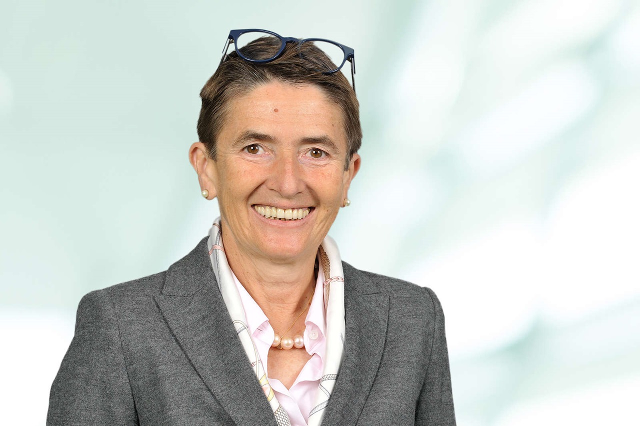 A person in a grey blazer and a light blouse with a pearl necklace. A blurred, bright background is present. Portrait of Zvezdana Seeger.
