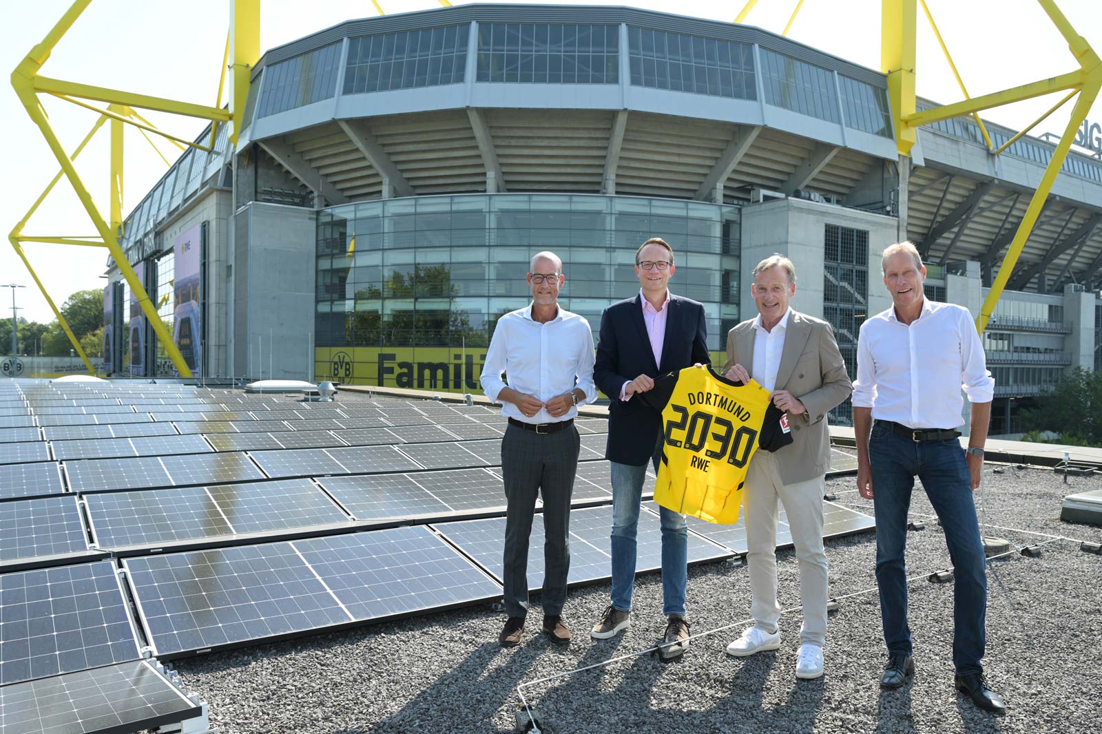 Four individuals stand on a rooftop covered with solar panels, holding a yellow jersey with 'Dortmund 2030 RWE' while a stadium looms behind.