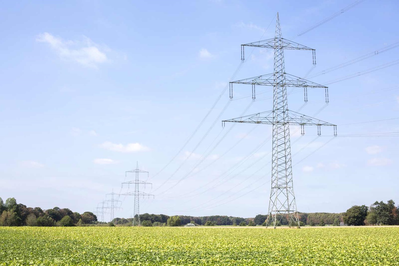 Tall electricity pylons rise above a vast green field under a clear sky with a few clouds.