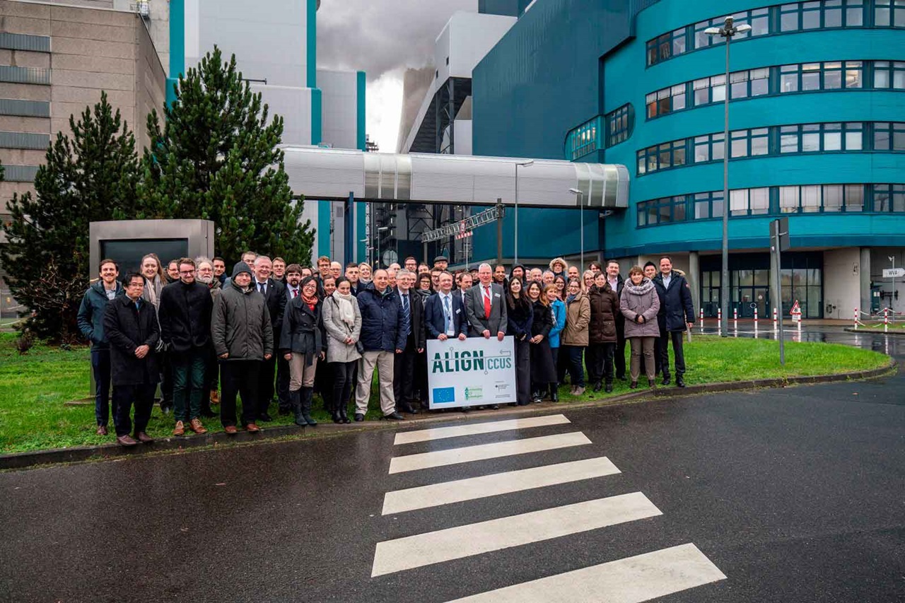Group photo of individuals in front of a modern building with greenery and a sign for the CCUS initiative.