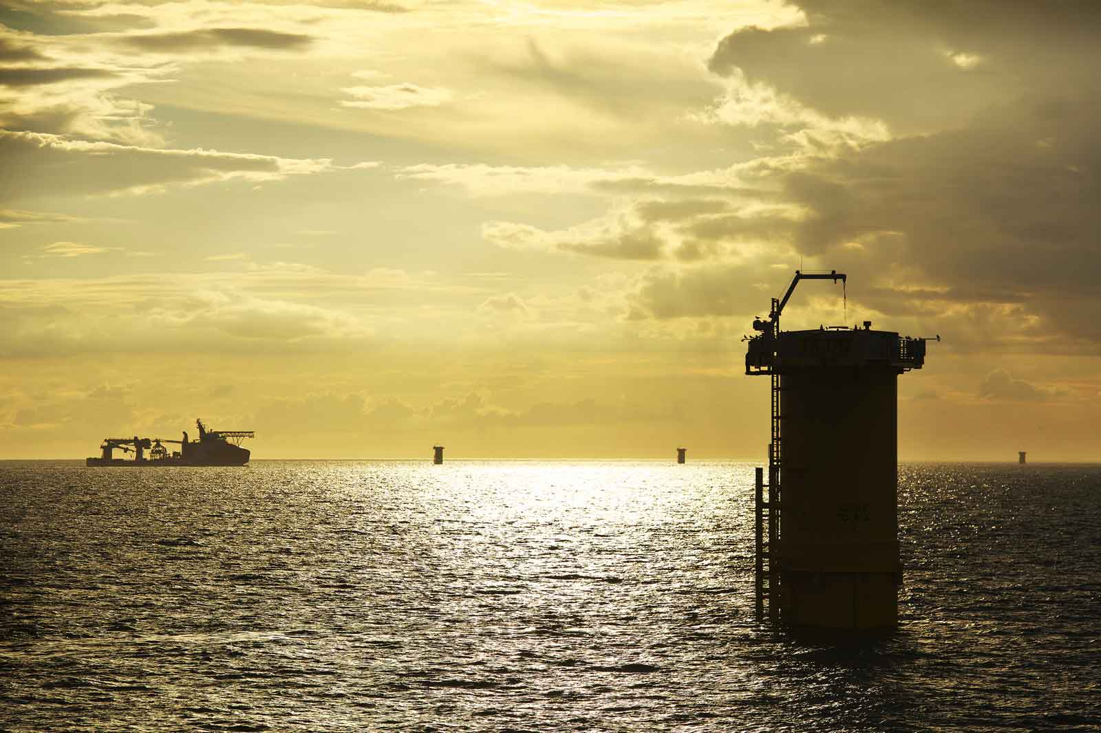 A platform stands in the water, with a silhouetted ship in the background during sunset.